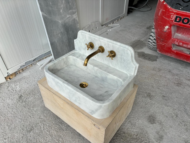Puede incluir: Un lavabo rectangular de m&aacute;rmol con grifer&iacute;a de lat&oacute;n, que incluye un grifo y dos manijas. El lavabo es de color gris claro con vetas grises m&aacute;s oscuras y se asienta sobre una base de madera de color claro. El grifo y las manijas tienen un acabado dorado.