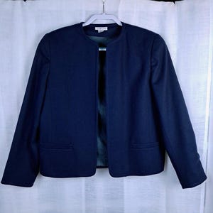 May include: A solid navy blue open-front jacket featuring a collarless round neckline, long sleeves, and two horizontal welt pockets at the lower front. The garment is displayed on a white plastic hanger and includes a visible interior brand label.