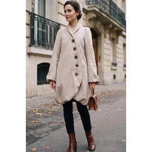 May include: A beige wool-blend coat featuring a shawl collar, long sleeves with gathered cuffs, and a single-breasted front closure with five dark buttons. The coat has a rounded, bubble-style hemline and is paired with dark trousers and brown boots.