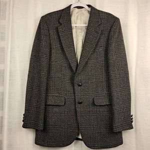 May include: A dark brown and grey plaid blazer featuring a notched lapel, two black front buttons, two flap pockets, and three smaller black buttons on each sleeve cuff. The interior is lined with a light-colored fabric and includes a white hanger.