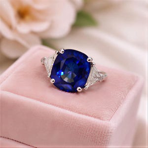 May include: A silver ring with a large, square-cut, deep blue gemstone as the centerpiece. Two smaller, triangular, clear gemstones flank the main stone. The ring is set on a pink velvet surface, with soft floral elements in the background.