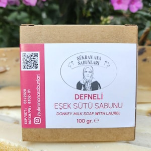 May include: A brown cardboard box with a pink and white label. The label features text in Turkish and English, including "Donkey Milk Soap with Laurel" and "100 gr. e". The box is placed in front of a blurred background of pink flowers.