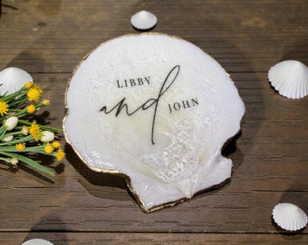 Può includere: Un piattino in resina bianco a forma di conchiglia con bordo dipinto in oro. Al centro è presente il testo stampato in nero "LIBBY and JOHN" in una combinazione di caratteri serif e corsivi. La superficie ha una finitura perlata e testurizzata.
