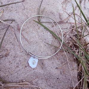 May include: A silver-toned bangle bracelet with a white, irregular-shaped charm. The charm has a small, silver-toned seashell design. The bracelet rests on a sandy surface with blades of grass.