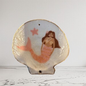 Peut inclure: Un vide-poche en forme de coquillage avec un motif de sirène peint. La sirène a des cheveux bruns, une queue rose et un collier de perles. Un bord doré et une étoile de mer rose ornent le fond blanc.