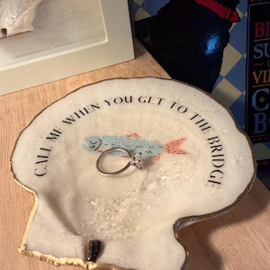 May include: A white, shell-shaped trinket dish with gold trim, featuring a light blue fish illustration and the text "CALL ME WHEN YOU GET TO THE BRIDGE." A silver ring with a clear stone rests on the dish.