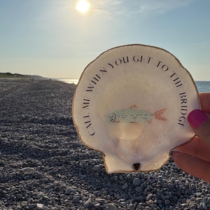 May include: A white scallop shell featuring a hand-painted illustration of a small fish in the center and the black text "CALL ME WHEN YOU GET TO THE BRIDGE" arched along the upper edge, with a metallic gold-painted rim around the shell's border.