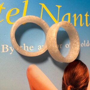 May include: Two circular rings with a smooth, semi-translucent off-white appearance featuring subtle speckled inclusions throughout the material. The bands have a uniform thickness and a rounded profile, displaying a consistent texture across their surfaces.