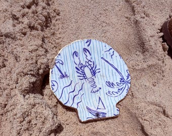Può includere: Un oggetto decorativo a forma di conchiglia con un design nautico blu e bianco con un'aragosta, una barca a vela e palme. I bordi sono rifiniti in oro. L'oggetto è appoggiato sulla sabbia.