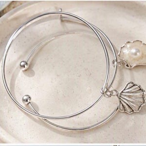 May include: A silver-toned bangle bracelet featuring two charms: one is a textured seashell shape and the other is a circular shell charm set with a small white pearl. The bracelet has a simple open design with two small silver spheres at the ends.