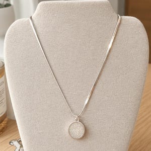 May include: A silver-toned necklace featuring a thin, flat snake chain and a circular pendant. The pendant has a smooth metallic rim surrounding a textured, shimmering white center that reflects light with a granular, glitter-like appearance.