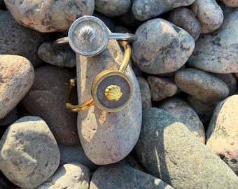 Può includere: Due bracciali aperti, uno color argento e uno color oro, ciascuno caratterizzato da un elemento centrale circolare con un inserto grigio testurizzato e un piccolo emblema a forma di conchiglia in rilievo abbinato al colore del metallo.