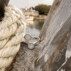 May include: A silver-toned open ring featuring two circular settings, each containing a textured, light gray, speckled inlay. The ring is positioned between a thick, twisted beige rope and a weathered, gray, textured stone surface.