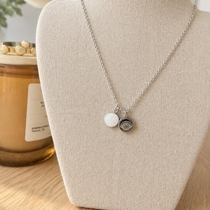 May include: A silver-toned chain necklace featuring two small circular charms. One charm has a solid white circular inlay, while the other is a dark circular disc engraved with a white line drawing of a seashell.