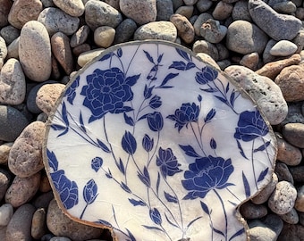 Può includere: Una conchiglia di capasanta con una superficie bianca decorata con un motivo floreale blu scuro di fiori in fiore e steli sottili. I bordi della conchiglia sono rifiniti con un bordo in metallo dorato e una piccola catena metallica è attaccata.