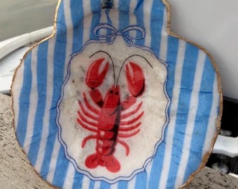 Può includere: Un piatto decorativo a forma di conchiglia di capasanta con un'illustrazione centrale di un'aragosta rossa all'interno di un bordo smerlato con un fiocco blu. Lo sfondo della conchiglia presenta strisce verticali azzurre e bianche, bordi dorati.