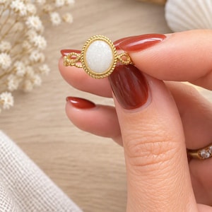 May include: A gold-toned ring featuring an oval-shaped white stone set within a twisted rope-style metal border. The band of the ring has a delicate, open-work twisted design, providing a textured appearance to the metal surface surrounding the stone.