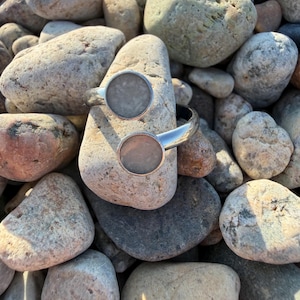 May include: A silver-toned open cuff bracelet featuring two circular ends, each inlaid with a smooth, light gray stone disc. The metal band has a polished finish and curves to create a bypass design, showcasing a clean and simple aesthetic.
