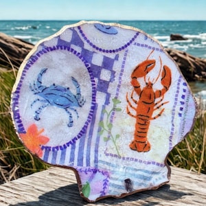 May include: A decorative seashell with a painted design featuring a blue crab and an orange lobster. The shell has a gold-colored edge and a patterned background with blue and purple accents. The shell is resting on a wooden surface.