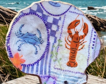 Può includere: Conchiglia decorativa con un disegno dipinto raffigurante un granchio blu e un'aragosta arancione. Il guscio ha un bordo color oro e uno sfondo a motivi con accenti blu e viola. Il guscio è appoggiato su una superficie di legno.
