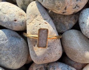 Può includere: Un anello dorato con una pietra rettangolare color beige chiaro incastonata in una semplice fascia metallica. L'anello è posizionato contro una collezione di ciottoli di fiume lisci e multicolori, evidenziando il contrasto tra il metallo lucido.