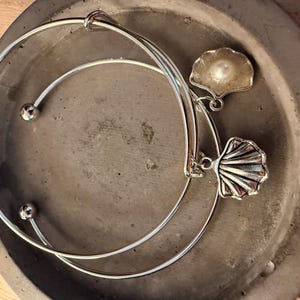 May include: Two silver-toned bangles with decorative seashell charms, one with a pearl accent, rest in a gray, circular dish. The bangles have a simple, elegant design, perfect for everyday wear. The seashell charms add a touch of coastal charm.