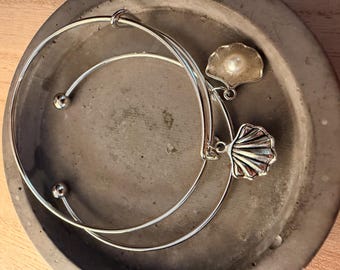 Può includere: Due braccialetti argentati con ciondoli a forma di conchiglia, uno con una perla, sono appoggiati in un piatto grigio circolare. I braccialetti hanno un design semplice. I ciondoli a conchiglia aggiungono un tocco costiero.
