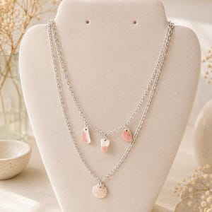 May include: A double-layered silver chain necklace featuring three small, irregular pink and white stone-like charms on the shorter top chain and a single round, textured pink seashell-style pendant on the longer bottom chain.