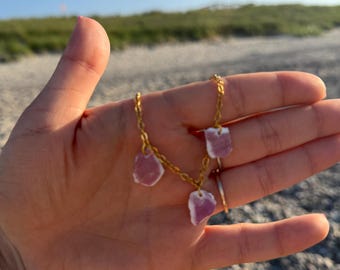 Può includere: Una collana a catena in metallo color oro con tre piccoli ciondoli in vetro marino rosa chiaro, traslucidi e di forma irregolare. I ciondoli sono distanziati lungo la catena, che presenta una delicata struttura a maglie.