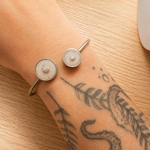 Puede incluir: Un brazalete abierto de tono plateado con dos extremos circulares. Cada extremo contiene una incrustación circular de color claro con un pequeño emblema de concha de tono plateado en relieve centrado en la superficie.