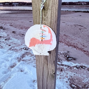 Könnte beinhalten: Ein weißes, muschelförmiges Ornament mit rosa und blauem Design, das an einem Holzpfosten hängt. Das Ornament zeigt einen Kartenumriss und farbenfrohe Akzente. Der Hintergrund zeigt eine Strandszene mit Schnee und Wasser.