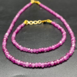Puede incluir: Un conjunto de collar y pulsera con pequeñas cuentas facetadas de color magenta. El collar y la pulsera tienen cierres dorados. Las joyas se exhiben sobre una superficie oscura.