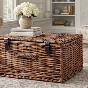 May include: A rectangular, woven wicker storage trunk with a hinged lid and leather straps. The trunk is brown and sits on a patterned rug. A vase of white flowers and books are on top. The trunk is 91 cm long, 51 cm wide, and 46 cm high.