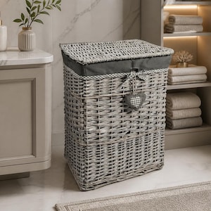 May include: A grey wicker laundry basket with a dark grey fabric lining and lid. The hamper features a heart-shaped charm. The basket is in a bathroom setting with a sink, towels, and a plant.