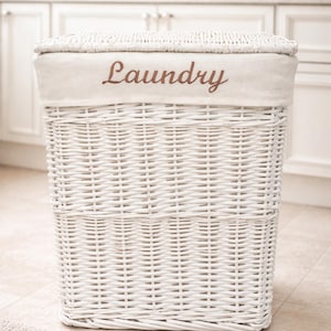May include: A white wicker laundry basket with a rectangular shape. The basket has a lid with the word "Laundry" embroidered in brown. The basket is on a light-coloured tiled floor.