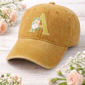May include: Mustard-colored baseball cap with a floral embroidered "A" initial. The cap features a curved brim and a fabric-covered button on top. The floral embroidery includes pink and white flowers with green leaves.