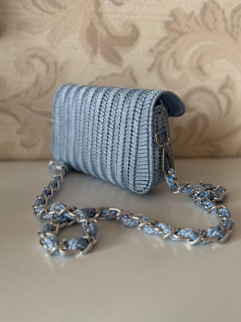 May include: A light blue woven crossbody bag with a silver chain strap. The bag is rectangular with a flap closure. The woven texture creates a subtle striped pattern. The chain is silver.