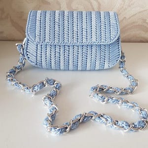 May include: A light blue crochet handbag with a silver chain strap. The bag has a rectangular shape with a flap closure. The crochet pattern features vertical stripes. The chain strap is a combination of silver metal and light blue crochet.