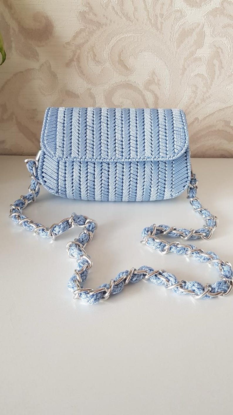 May include: A light blue crochet handbag with a silver chain strap. The bag is rectangular with a flap closure. The crochet pattern has vertical stripes. The chain strap combines silver metal and light blue crochet.