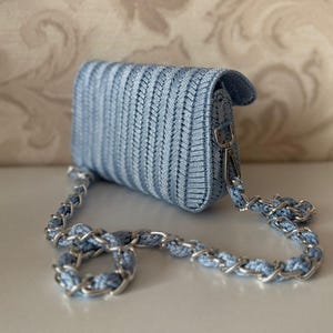 May include: A pale blue woven crossbody bag with a silver chain strap. The bag features a flap closure and a textured design. The chain is a combination of silver links and pale blue woven material. The bag is on a white surface.