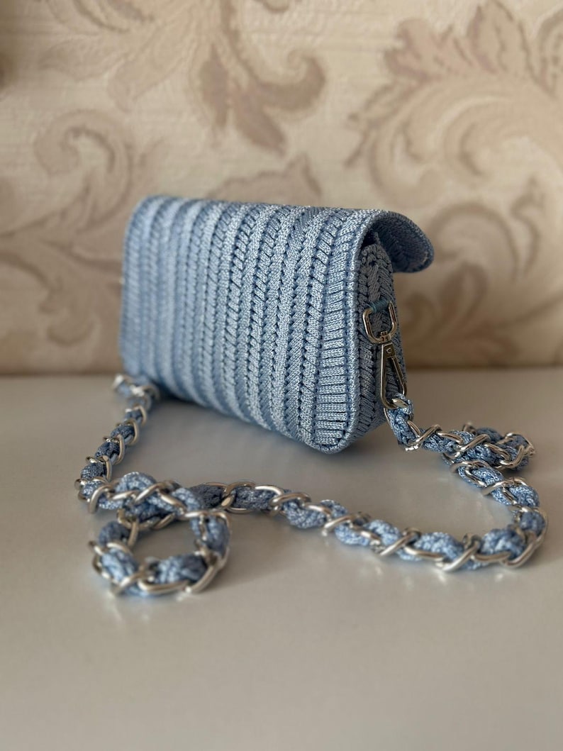 May include: A pale blue woven crossbody bag with a silver chain strap. The bag features a flap closure and a textured design. The chain is a combination of silver links and pale blue woven material. The bag is on a white surface.