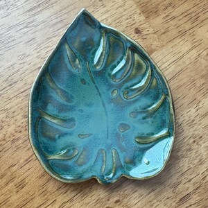 Może przedstawiać: Ceramiczna miseczka w kształcie liścia monstery z błyszczącym wykończeniem. Naczynie pokryte jest szkliwem w odcieniach głębokiego turkusu i zieleni, z ciemniejszymi akcentami podkreślającymi żyłkowanie oraz krawędzie liścia.