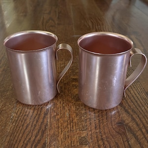 May include: Two cylindrical copper mugs with smooth, metallic surfaces and curved handles. Each mug features a polished finish with subtle variations in tone, showcasing the distinct reddish-brown color of the metal. The mugs are open at the top.