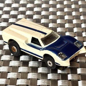 May include: A vintage toy race car in cream and blue. The car has a cream-colored body with a blue hood and racing stripes. The wheels are black with a gold center. The car is a miniature replica of a classic sports car.