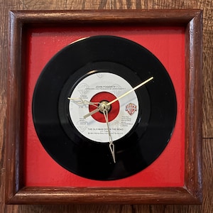 May include: A square clock with a wooden frame and a red background. The clock face is a black vinyl record with gold hands. The record label reads "JOHN FOGERTY" and "THE OLD MAN DOWN THE ROAD".
