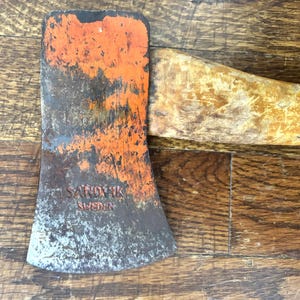 May include: Vintage axe with a weathered orange head and wooden handle. The axe head has the text "SANDVIK SWEDEN" in red. The axe is set against a wooden background.