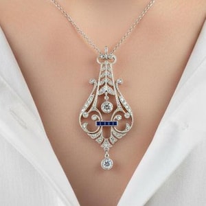 May include: A silver-toned pendant necklace featuring an intricate, elongated filigree design adorned with small clear stones and a horizontal row of three deep blue rectangular stones, suspended from a delicate silver-toned chain.