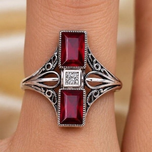 May include: A silver ring featuring two rectangular deep red gemstones positioned vertically with a small square white gemstone set between them. The band displays intricate metalwork with open scroll patterns on both sides of the central stones.
