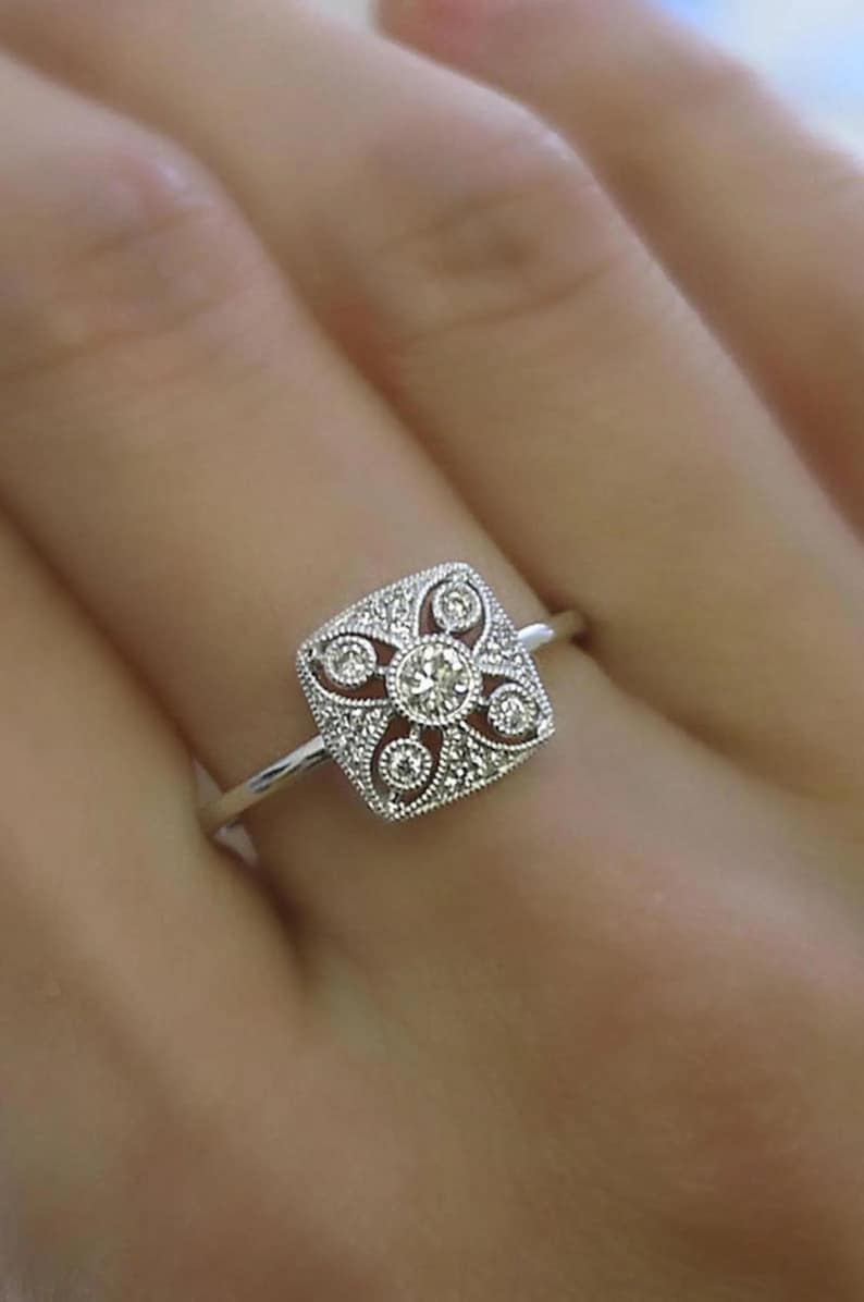 K&ouml;nnte beinhalten: Ein silberner Ring mit einem quadratischen, verzierten Gesicht. Das Gesicht hat einen zentralen runden Diamanten, der von kleineren Diamanten und dekorativen Filigranarbeiten umgeben ist. Der Ring hat ein d&uuml;nnes, poliertes Band.