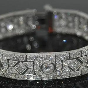 May include: A silver-toned metal bracelet featuring a repeating pattern of circular and geometric links. The entire surface is encrusted with numerous small, clear, sparkling stones set in a dense, intricate arrangement across the band.